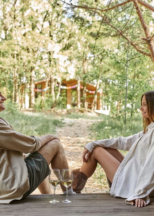 Vivre et relaxer sur un patio conçu sur mesure – Patio Exotic Homme et femme assis sur une terrasse en bois, dégustant du vin, avec un gazebo en arrière-plan, entourés de verdure, illustrant un espace de vie extérieur confortable et accueillant.
