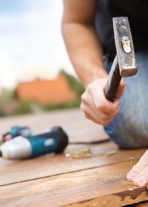 Pose de bois pour patio – Conception Patio Exotic Homme utilisant un marteau pour fixer une vis sur un patio en bois, avec une perceuse à proximité, illustrant le processus de construction de patios personnalisés par Patio Exotic.