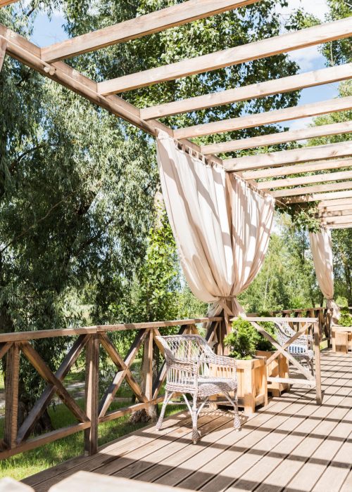 Patio avec pergola et rideaux – Design Patio Exotic Patio en bois avec pergola et rideaux, chaises confortables et verdure environnante, représentant un espace extérieur élégant et accueillant pour un aménagement personnalisé.