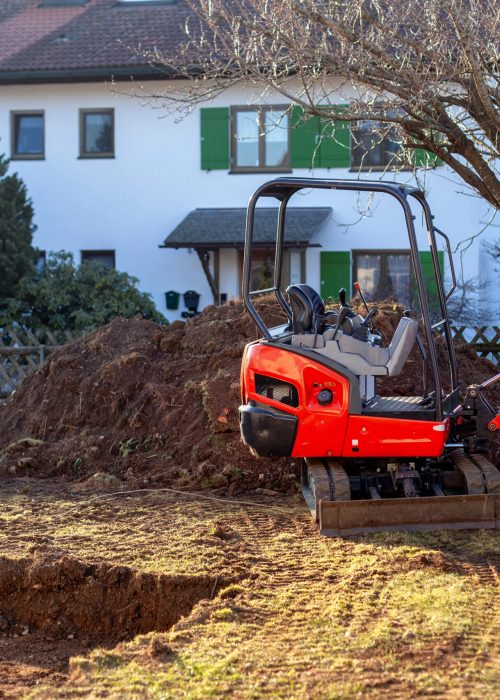 Mini-excavatrice rouge sur un site d'excavation résidentielle, avec un tas de terre en arrière-plan, illustrant les services d'aménagement extérieur de Patio Exotic.