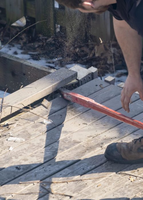 Homme utilisant un outil pour démolir un vieux patio, avec éclats de bois et débris sur le sol, illustrant le service de démolition de Patio Exotic.