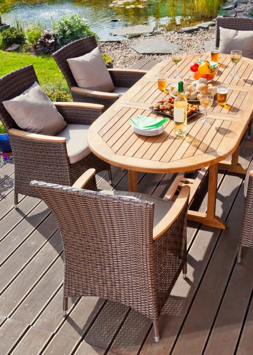 Table en bois avec chaises en rotin sur une terrasse, décorée de fruits, boissons et collations, entourée d'un jardin verdoyant et d'un étang, illustrant un espace extérieur accueillant pour des repas en plein air.
