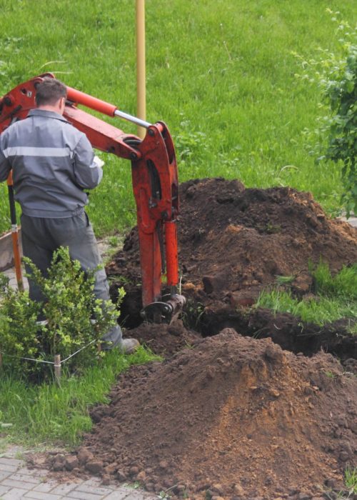 Ouvrier utilisant une mini-pelle pour effectuer des travaux de terrassement résidentiel dans un jardin, avec des tas de terre et une pelouse verte environnante.