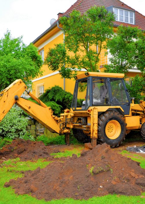 Excavatrice jaune en action dans un jardin résidentiel, creusant un sol pour des travaux d'aménagement extérieur.