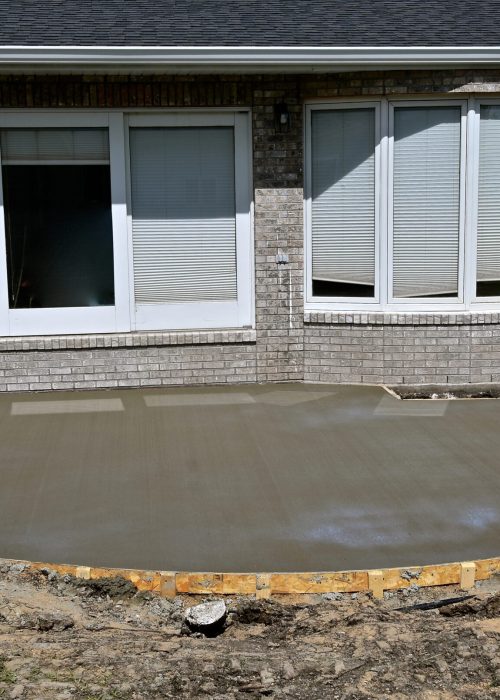 Fondation de patio en béton fraîchement coulée, montrant une surface lisse et des bordures en bois, avec une maison en briques à l'arrière-plan.