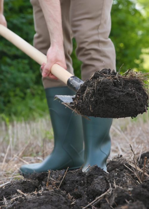 Homme en bottes en caoutchouc utilisant une pelle pour manipuler de la terre, illustrant les services de terrassement résidentiels de Patio Exotic.