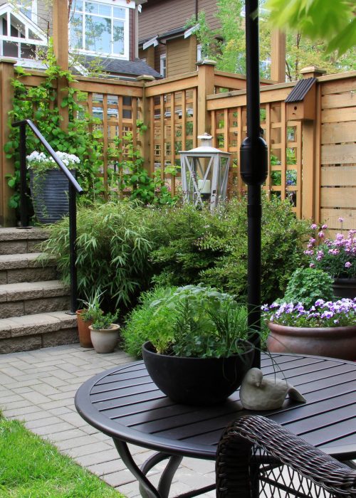 Espace extérieur accueillant avec patio en pierre, table ronde en métal, plantes en pot, clôture en bois, et lanternes, illustrant l'expertise de Patio Exotic en aménagement de jardins et installations de clôtures sur mesure.
