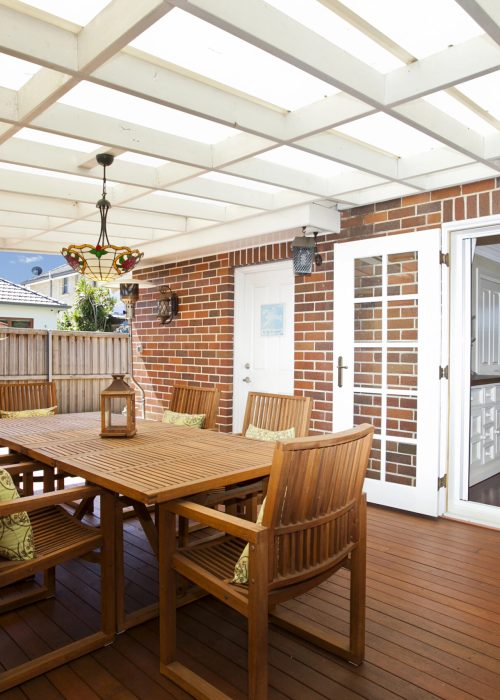 Terrasse en bois avec table et chaises, éclairage suspendu, et porte d'entrée, illustrant un espace de vie extérieur esthétique et fonctionnel pour Patio Exotic.